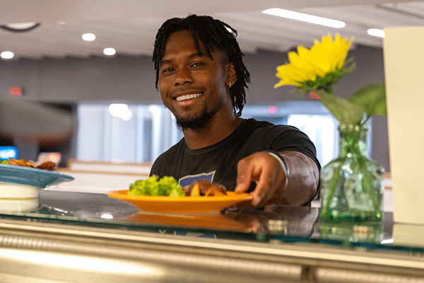 An African-American male students smiles while being handing a plate of food in the dining hall of Sycamore Towers.